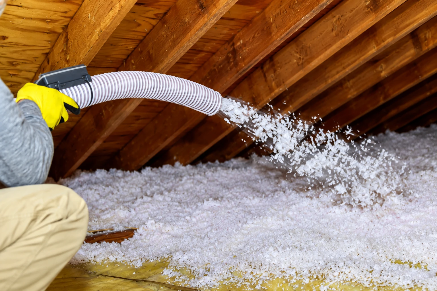A worker directs a hose to blow loose-fill insulation across an attic floor beneath exposed roof rafters.