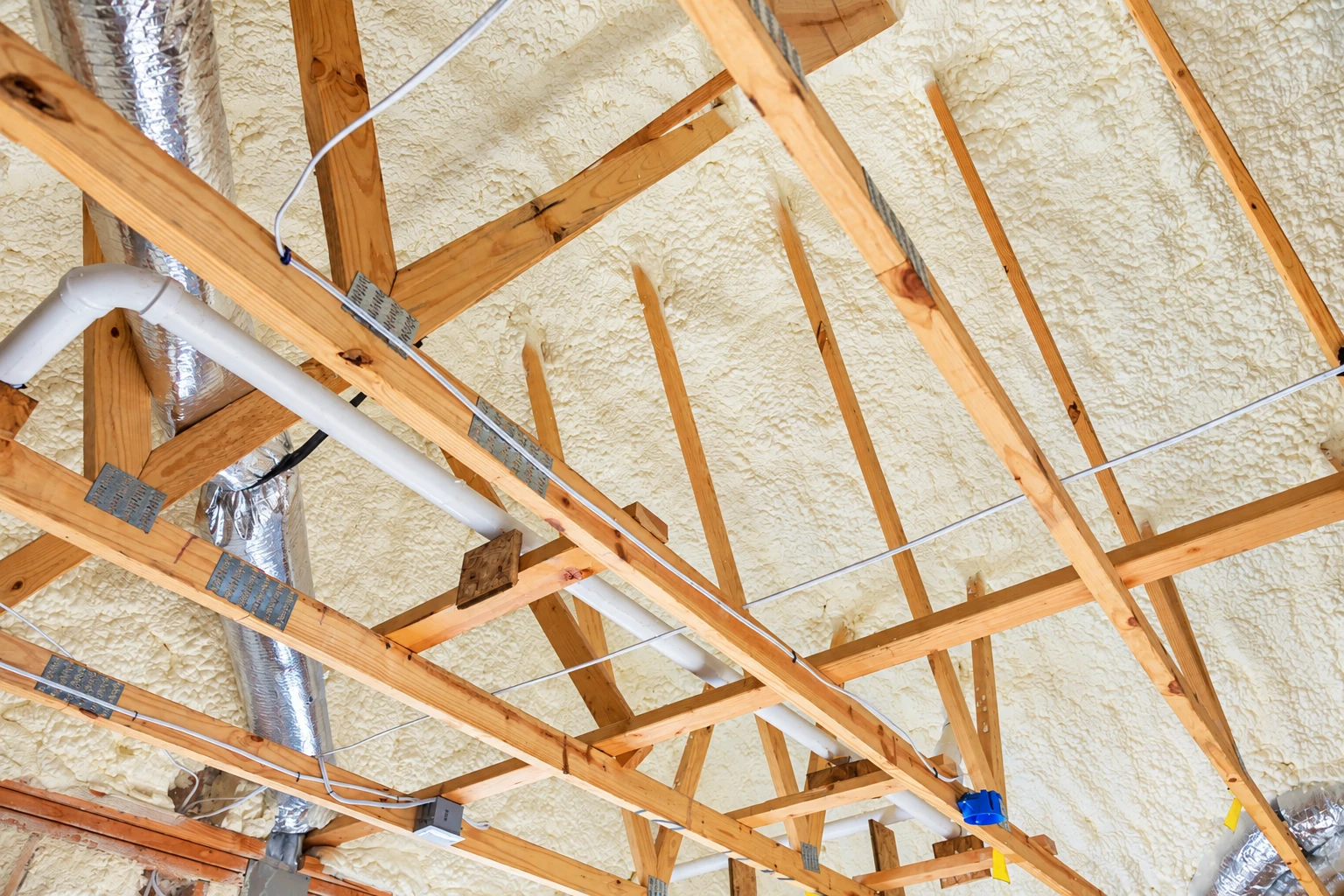 Closed-cell spray foam covers the underside of a roof among exposed wooden trusses, pipes, and wiring.