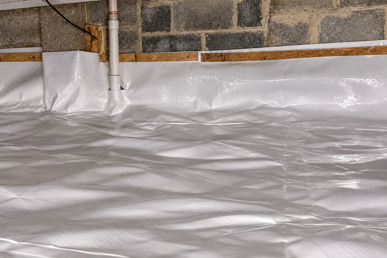 A white plastic vapor barrier covers the floor and lower walls of a stone foundation crawl space.
