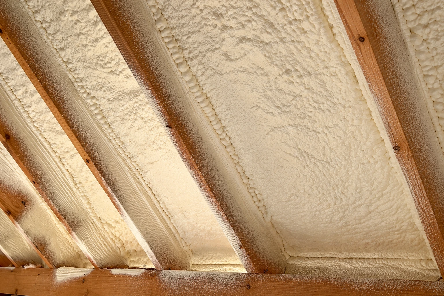 Cream-colored spray foam fills the spaces between exposed wooden roof rafters.