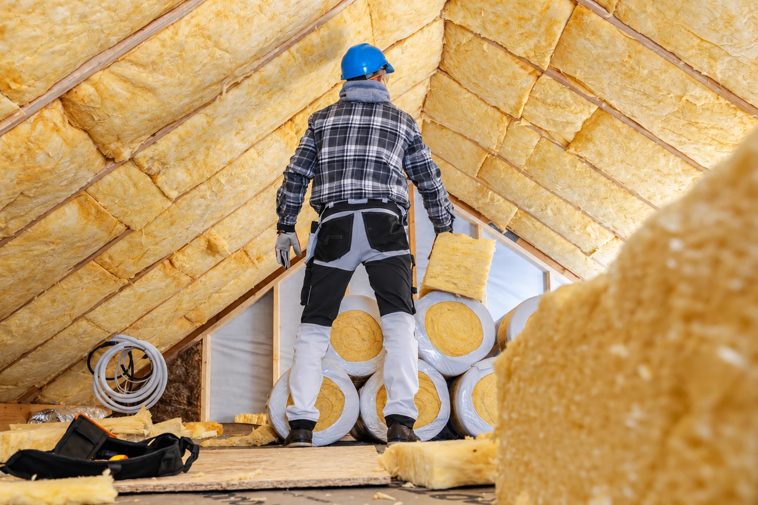 A worker in a blue hard hat stands among yellow insulation rolls in an attic lined with fiberglass batts.