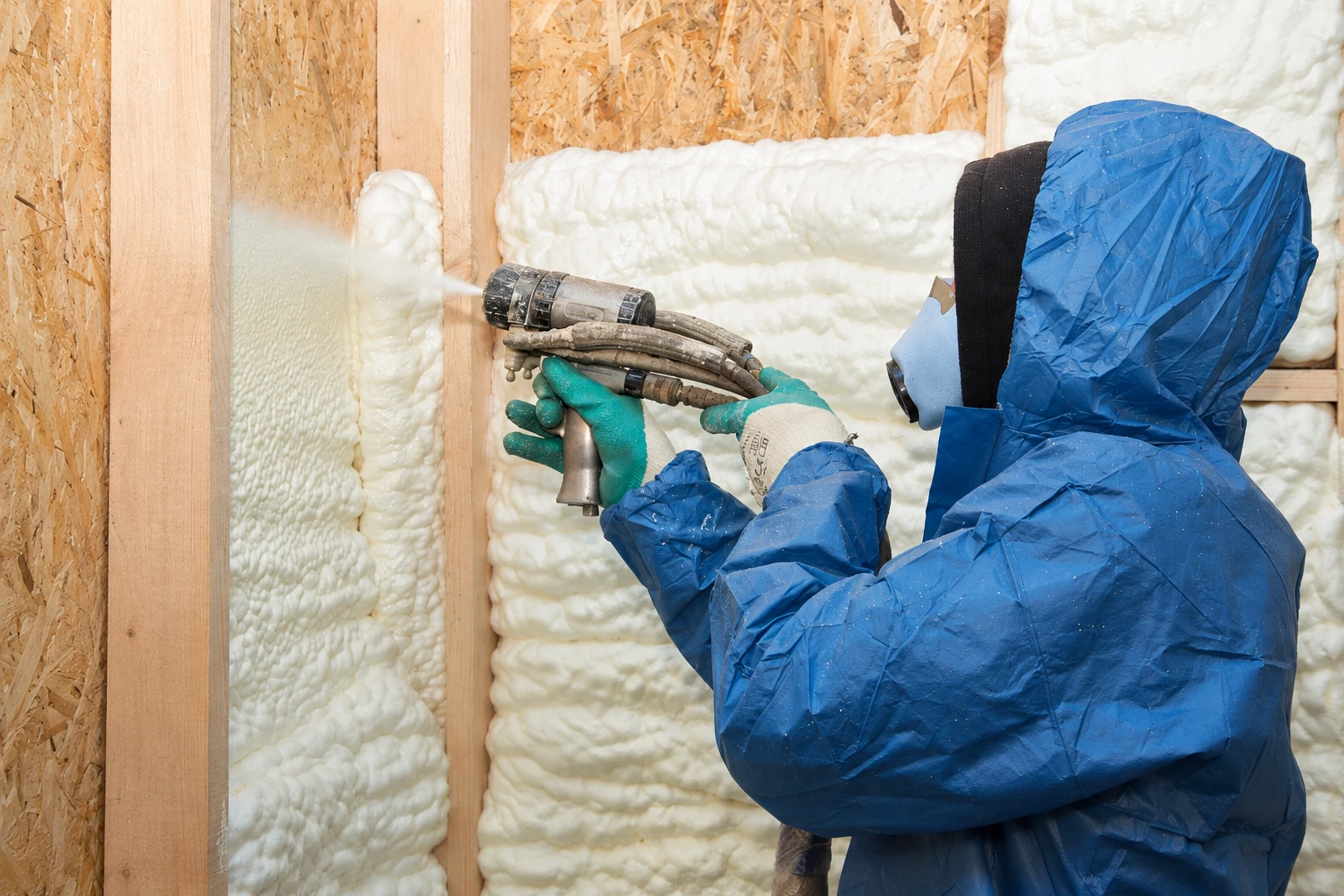Worker in protective gear sprays white foam insulation into the cavities of a framed wall.