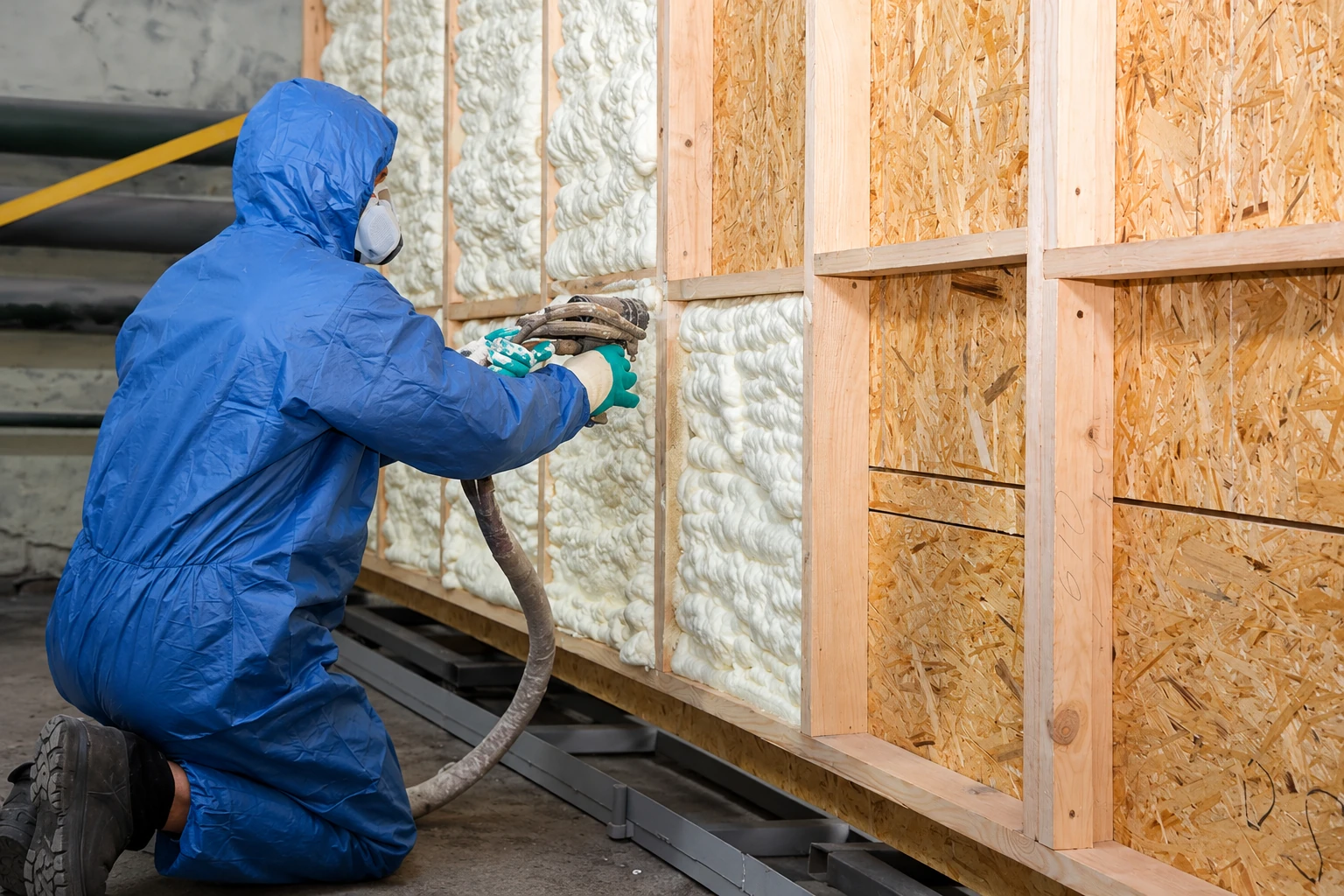 A worker in blue protective coveralls sprays foam insulation into open wall studs.